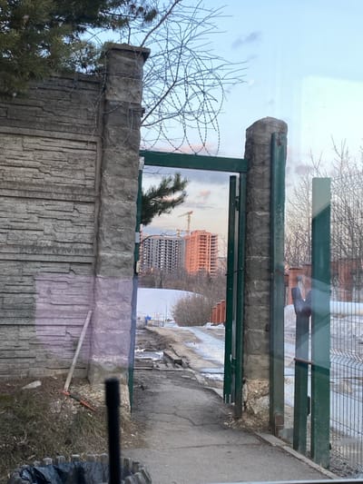 This image, located in Марфино, Russia, depicts a scene through a partially open green metal gate and an adjacent concrete wall. The foreground shows a black cylindrical object in the lower center, and part of a wooden planter containing a black trash bag in the lower left. Reflections are visible on the surface from which the photograph was taken, suggesting a glass barrier.
On the left, a textured grey concrete wall stands, adjoined by a substantial concrete gate post topped with coils of barbed wire. A green pine branch extends from above the wall into the frame. Leaning against the wall near its base is a tool resembling a shovel or rake with a red handle.
The open gate, constructed of green metal bars and supported by a second concrete post, reveals a downward-sloping, cracked asphalt path covered in patches of mud, dirt, and melting snow. Bare deciduous trees and shrubs line the path beyond the gate. To the far right, a segment of a metal wire fence is visible.
In the mid-distance, a landscape of sparse winter vegetation and patches of snow leads to a construction site. Two multi-story buildings are under construction: one grey and incomplete, and another reddish-orange brick structure, illuminated by warm, possibly sunset light. A yellow tower crane is positioned between these buildings. The sky above is partially cloudy, displaying shades of blue, pink, and orange. No individuals are present in the scene.