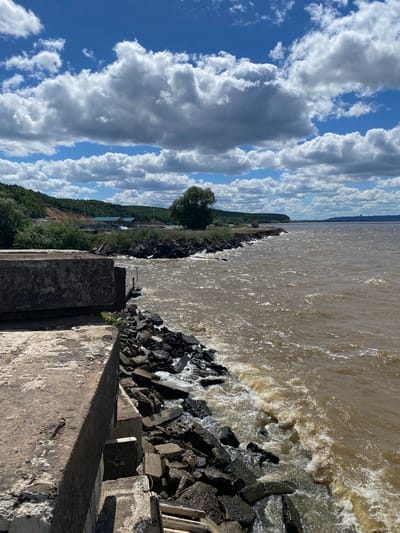 The image depicts a view of the Volga River in Leninsky, Russia, on a cloudy day. The foreground features a weathered concrete structure, possibly a pier or embankment, with visible rebar. Large rocks and rubble form a jagged edge along the water's interface with the concrete. The river itself is a muddy brown color, with choppy waves and white foam indicating some turbulence. A rocky shoreline extends into the distance, bordered by lush green vegetation and a gently sloping hill covered in trees. Along the distant shoreline, there are several buildings with light-colored roofs and a few masts, suggesting a small settlement or harbor. The sky is a mix of bright blue patches and large, white cumulus clouds, some of which cast shadows. The overall scene is natural and somewhat rugged, with a sense of expansive water and a clear, albeit partially overcast, sky.