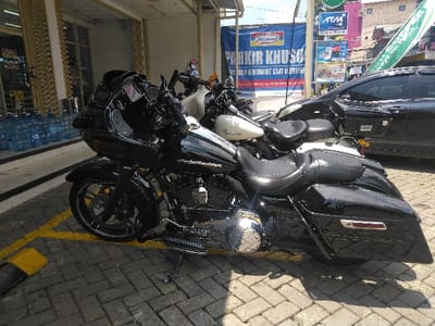 A black Harley-Davidson touring motorcycle, featuring a large front fairing, chrome engine details, and black hard saddlebags, is parked on an interlocking brick pavement. The motorcycle is positioned on its kickstand, occupying a space marked by yellow painted lines and a yellow-and-black parking block. Behind it, a white motorcycle is partially visible, and further right, a black sedan or SUV is parked. To the left, a storefront with large glass windows reveals stacks of large blue plastic water dispenser bottles. The background features a large blue banner prominently displaying "INDOMARET" and "PARKIR KHUSUS" (Indonesian for "SPECIAL PARKING"), along with other advertising signs including an "ATM" logo. The visual evidence, particularly the Indomaret branding and Indonesian text, indicates the scene is located in Indonesia, contradicting the provided location of La Roche-en-Ardenne, Belgium. No individuals are visible.