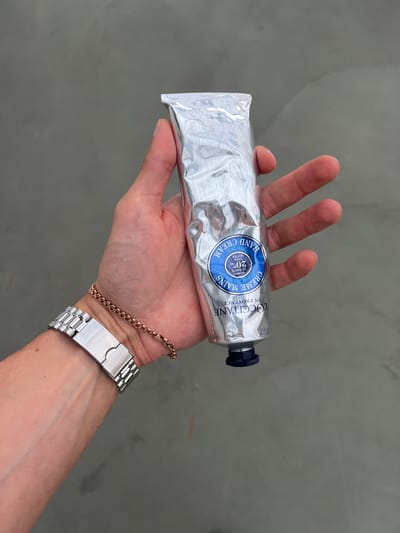 An individual's hand is holding a silver tube of L'Occitane Shea Butter Hand Cream. The hand is adorned with a silver watch and a thin rose gold bracelet. The background is a plain, gray, textured surface, possibly concrete, with subtle lighting variations. The image does not provide any information about the city of Borowa, Poland.