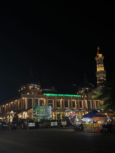 This is a nighttime photograph of the Grand Mosque of Darul Muttaqin in Tanggul, Indonesia. The mosque features prominent domes and a tall minaret, both illuminated by artificial lights. In front of the mosque, a street is lined with parked cars and several food stalls. One stall prominently advertises "Spesial Siomay & Cilok." A banner on the mosque's facade reads "Takmir Masjid Besar Darul Muttaqin Tanggul" and congratulates the community on the Islamic New Year 1448 Hijriah, also mentioning a charitable event for orphans. The scene is vibrant with the glow of lights and the activity of street vendors and passersby.