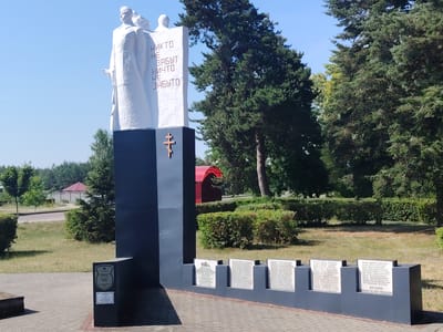 This is a photograph of a war memorial located in Malaryta, Belarus. The memorial features a tall, black, rectangular base topped with white sculptures of three figures, two of whom are visible in full. The white sculptures are inscribed with Cyrillic text that translates to "No one is forgotten, nothing is forgotten." A small wooden cross is affixed to the front of the black base.
At the foot of the memorial, a dark gray, stepped wall displays several plaques inscribed with lists of names in Cyrillic. The largest plaque at the far right reads "ВЕЧНАЯ ПАМЯТЬ ПОГИБШИМ," meaning "Eternal memory to the fallen." Another plaque on the left side of the base shows a shield emblem and some text.
The memorial is situated outdoors in a park-like setting. Lush green trees, manicured bushes, and a paved walkway surround it. In the background, a red-roofed structure and a fence with trees beyond suggest a developed area. The sky is a clear, bright blue, indicating a sunny day. The overall atmosphere is solemn and commemorative, likely marking an event of historical significance related to war and remembrance.