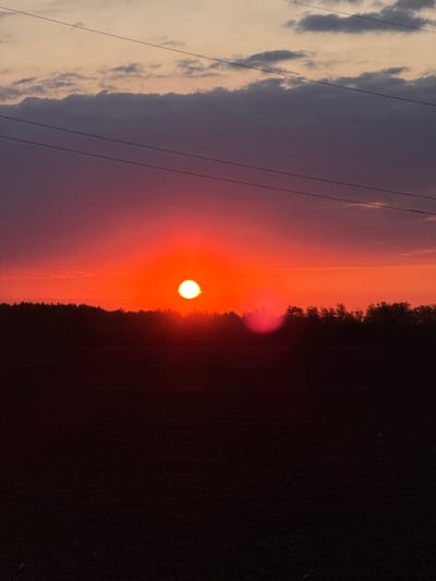 A landscape-oriented image depicts a sunset or sunrise. The left side of the frame contains clouds, with the sky transitioning to shades of pink and orange. The sun is a bright white circle. The right side features a dark, silhouetted treeline. A set of power lines is visible across the sky.

It is not possible to determine the exact location in Bavly, Russia, based solely on the provided image.