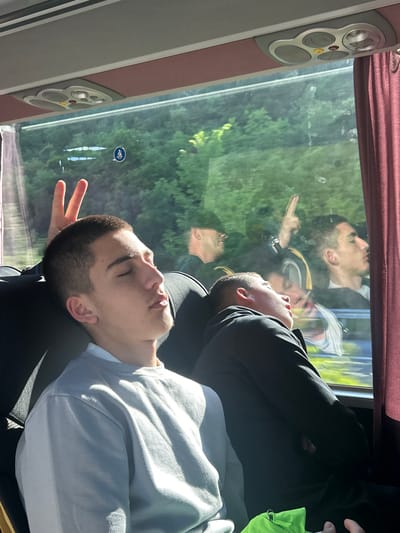 A young man sleeps against a seat on a bus or train. His head leans against the headrest. He wears a light-colored sweatshirt. Another man is sleeping behind him, wearing a dark jacket. The window shows a reflection of people outside, as well as a view of trees and vegetation. The interior of the bus or train is visible, with window trim and controls. There is a blue handicap symbol on the window. Without additional information, it is impossible to confirm that the image was taken in the Municipal Unit of Kato Olympos, Greece.