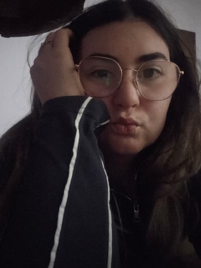 The image displays a close-up portrait of an individual, purportedly located in Negorci, North Macedonia, within an indeterminate indoor setting. The subject is a young adult female with dark, long hair, wearing large, round, gold-rimmed eyeglasses. Her lips are pursed, and she is looking directly at the camera. Her right hand is visible, resting against the right side of her head, with a ring on her right index finger. She is wearing a dark, long-sleeved garment, possibly a track jacket or sweatshirt, featuring a vertical white stripe down the outer seam of the left sleeve and a visible zipper pull on the front. The background is largely out of focus, appearing as a light-colored surface behind her upper left shoulder and darker, indistinct areas elsewhere. The lighting appears dim.