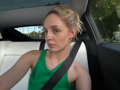 A young woman with blonde hair pulled into a messy bun is in the passenger seat of a car. She is wearing a bright green tank top and a gold necklace. A black seatbelt is across her chest. The interior of the car is visible, with white leather seats and a dark dashboard. Outside the car, through the windows, palm trees and houses are visible under a bright sky, suggesting a location in Coral Gables, United States of America, likely during the daytime. The woman is looking towards the camera with a neutral expression.