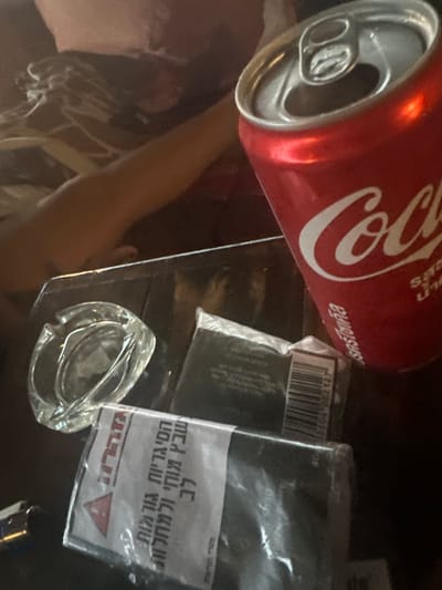 The image displays an indoor scene in the city of Ban Paem Bok, Thailand, centered on a dark, reflective surface, likely a table. A red Coca-Cola can, open with its silver tab visible, occupies the upper right portion of the frame and displays Thai script. To the left of the can, a clear glass ashtray rests on the surface, appearing empty. Adjacent to the ashtray is a black, rectangular packaged item, partially wrapped in plastic, with a visible barcode and text. Below this item and towards the bottom left, a white-labeled package, possibly containing cigarettes, is visible. This package features a red warning triangle and text in Hebrew, including the word "אזהרה" (Warning). A metallic lighter is partially visible in the bottom left corner. In the blurred background, a bare human arm and a portion of a torso are discernible in the upper left.