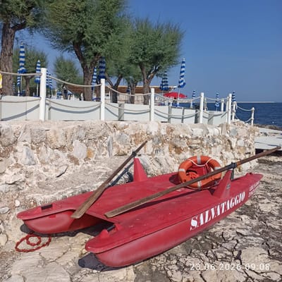 The image captures a bright, sunny day at a beach in Molfetta, Italy. In the foreground, a red, double-hulled inflatable boat, labeled "SALVATAGGIO" (meaning "Rescue" in Italian), rests on a rocky shore. Two wooden oars are laid across the boat, and an orange life preserver with a rope is visible. The boat appears to be a rescue vessel, possibly for a beach concession.
Behind the boat, a low wall constructed of rough, light-colored stones separates the rocky beach from a raised area. This raised area features a white railing with rope accents, separating a walkway or lounge area from the beach. Several closed beach umbrellas with blue and white stripes are arranged behind the railing, along with some blue beach chairs and a small wooden structure, possibly a lifeguard station or kiosk. A bright red umbrella is also visible further down the beach.
In the background, the blue expanse of the Adriatic Sea stretches to the horizon, with a distant breakwater or pier visible. Lush green trees with fine foliage, likely tamarisks, line the area behind the beach infrastructure. The sky is a clear, deep blue. The overall scene depicts a well-maintained beach facility on a calm day. A timestamp in the lower right corner reads "23.06.2026 09:08."