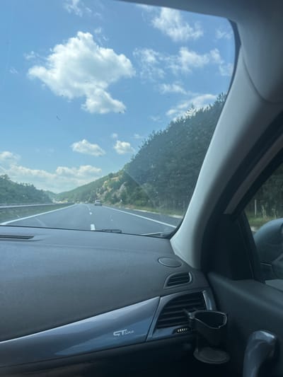The image shows a view from inside a car looking out at a highway in a rural area. The road is paved and has white lines marking the lanes. There are trees and hills on either side of the road, and the sky is blue with some white clouds. The dashboard of the car is visible in the foreground, with a "GT Line" logo on the lower part. There is a car holder attached to the air vent. The location is the highway near Dolno Varshilo, Bulgaria.