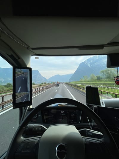 The image is a view from the driver's seat of a Volvo truck traveling on a highway near Mezzocorona, Italy. The speedometer reads 82. Mountains are visible in the distance. A monitor in the cab shows rear views. A roll of paper towels is hanging from the cab's ceiling.