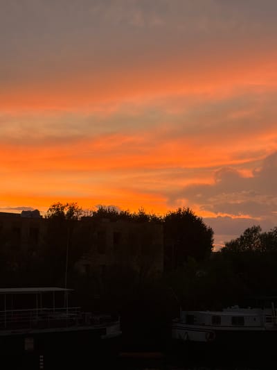 The image captures a vibrant sunset over the city of Vienne, France. The sky is a dramatic gradient of fiery orange and soft grey, with streaks of pink and yellow illuminating the clouds. In the foreground, silhouetted against the dramatic sky, are the dark shapes of trees and buildings. Two boats are visible on the water, one on the left with a covered deck and railings, and another on the right, a smaller vessel with windows. The overall scene evokes a sense of tranquility and natural beauty.