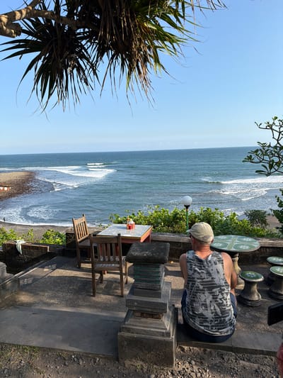 A man wearing a camouflage tank top and a tan baseball cap sits on a stone ledge, facing away from the camera, looking out at the ocean. He is at an outdoor dining area with a table and chairs in the foreground. The table has a checkered tablecloth and a bottle of red sauce on it. Beyond the table and chairs is the ocean, with waves breaking on the shore. In the background, a beach stretches along the coastline. The area is covered in stone pavers and there is lush green vegetation around the seating area. The scene appears to be in a restaurant or cafe with an ocean view in Selemadeg Barat, Indonesia.