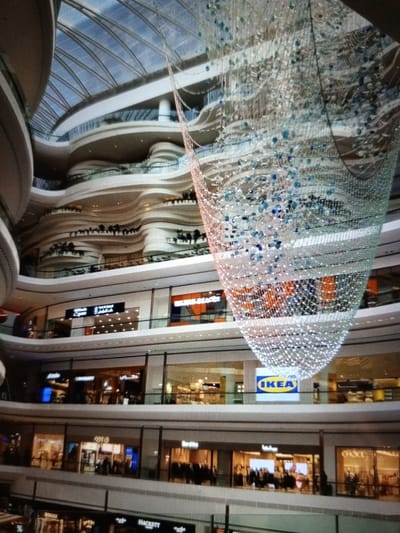 The image depicts the interior of a multi-level shopping mall with a large, ornate chandelier hanging from the ceiling. The architecture features curved balconies and a transparent roof. Several shops are visible on different levels, including one with an IKEA sign. People are present, browsing and walking within the mall. The environment appears to be clean and well-lit, typical of a modern commercial space. The location is identified as Golbembal, Nigeria.