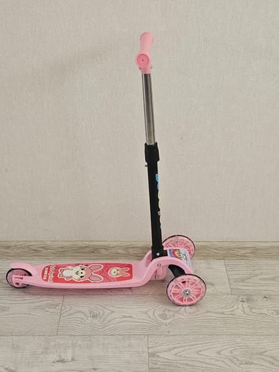A pink three-wheeled scooter stands upright on a light-colored wooden floor against a textured beige wall. The scooter has a silver adjustable handle with pink grips. The platform of the scooter is also pink and features a graphic of two cartoon bunnies. The wheels are pink and translucent with silver hubs. The scene is indoors, likely a home environment, and there is no indication of any specific location in the city of Jezqazğan, Kazakhstan.