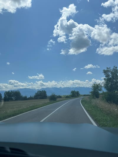 A wide shot captures a scenic view of a paved road winding through a rural landscape under a bright blue sky dotted with fluffy white clouds. The road, marked with white lines, curves gently to the right, disappearing into the distance. To the left of the road, a field of dry, golden grass stretches towards a line of trees. Beyond the trees, a range of hazy blue mountains forms the horizon. To the right of the road, tall grass and green foliage grow, with a large, leafy tree dominating the right edge of the frame. The foreground shows the dark dashboard of a vehicle, suggesting the photo was taken from a car. The scene evokes a sense of travel and natural beauty, with no people visible. The location is identified as a road in or near the city of Voila, Romania.