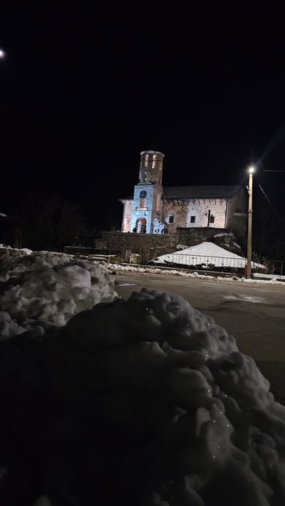 The image presents a night scene in Sitovo, Bulgaria, characterized by winter conditions and artificial illumination. In the immediate foreground, large, irregular mounds of snow dominate the lower left and extend partially across the frame. These snow piles exhibit sparkling surfaces from reflected light. An asphalt road, partially covered with snow and slush, recedes into the midground. To the right of the road, a low, dark metal fence borders a snow-covered mound. A tall utility pole with a bright streetlamp stands on the far right, casting a strong beam of light.
The central subject is a stone church situated on elevated ground behind the road. The church features a multi-tiered bell tower on its left, with visible arched openings illuminated by warm, amber light from within. A clock face is discernible on the tower. The main facade, including an arched entrance, is brightly illuminated with blue light. The church is constructed of stone masonry, with a dark roof. A dark cross is positioned on the roofline to the right of the bell tower. The structure is supported by a stone retaining wall. Bare trees are visible against the dark sky on the left side, behind the church.
The sky is dark, indicating nighttime. A distinct crescent moon is visible in the upper left corner of the frame, and a faint point of light, possibly a star, appears in the upper right. No individuals or discernible activities are present in the scene.