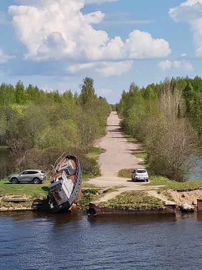 The image depicts a scene in Ivanov Bor, Russia. An old, damaged ship is partially submerged at the water's edge. A silver SUV is parked on the grassy bank behind the ship, and a white compact car is parked across the road. A paved road stretches into the distance, lined on both sides by trees. The sky is blue with scattered white clouds.