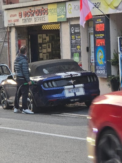 A vertical shot of a gas station showing a man fueling a blue and white striped sports car. The gas station sign reads "eectrauto PROXIMEGA," with "TOKSNES STATION SERVICE" underneath. Several other signs indicate "ROMANO ENERGY," with displayed fuel options and prices. The sports car, identifiable by the license plate "4895", is parked next to a red vehicle with visible tires. The scene captures a typical gas station activity in a somewhat vertical orientation.