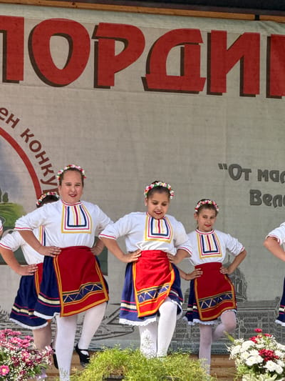 The image depicts a group of young girls in traditional Bulgarian folk costumes performing on a stage, likely outdoors, in Pordim, Bulgaria. The backdrop features a large banner with prominent red lettering, parts of which are visible and read "ПОРАДИ" and "ЕН КОНКУ". The girls, adorned with floral headbands, are wearing white blouses with colorful embroidered collars and red skirts over dark blue underskirts. They are positioned in a row, some with hands on their hips, suggesting a choreographed dance. In the foreground, there are floral arrangements and greenery, indicating a festive event. The overall impression is one of a cultural performance or festival in the town of Pordim.