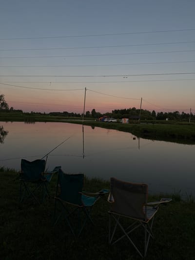 The image depicts a serene waterfront scene at dusk, likely in Komunar, Russia. The foreground is dominated by two dark green camping chairs positioned on grassy terrain. One chair is closer to the camera and slightly angled, while the other is further back and more upright. Beyond the chairs, a calm body of water reflects the dusky sky and the surrounding landscape.
In the middle ground, a thin fishing rod is extended over the water, suggesting an angling activity. Further in the background, a line of trees forms a dark silhouette against the horizon. Among the trees, several tents and a white vehicle are visible, indicating a campsite or a gathering. A tall utility pole with multiple horizontal wires stretches across the frame, dividing the sky.
The sky exhibits a gradient of colors, transitioning from a pale blue at the top to soft pink and orange hues near the horizon, characteristic of sunset or twilight. The overall mood is peaceful and tranquil, with the stillness of the water and the fading light contributing to a sense of calm. No visible text is present in the image.