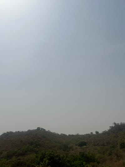 The image displays a panoramic landscape under a hazy, light blue-grey sky that occupies approximately the upper two-thirds of the frame. The sky shows a slightly brighter luminosity towards the upper left, indicative of a obscured sun. The lower third of the image features undulating hills and slopes covered with dry, sparse vegetation, predominantly brown and dull green scrubland, suggesting a dry season. Scattered across the terrain are several trees, with a more prominent, denser green tree situated within a valley-like depression in the midground. Rocky formations are visible atop some of the hills in the background. No human figures, man-made structures, or animals are visible within the scene. The environment appears natural and undeveloped. This scene is located in Kwakwa Laji, Nigeria.