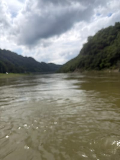 The image depicts a blurry, wide-angle view of a body of water, likely a river, with forested hills on either side. The water has a greenish-brown hue, and there are reflections of light on its surface, appearing as small, bright circles. The sky is overcast with dark, dramatic clouds. The scene suggests a natural, possibly remote, environment. The provided location of Seo-myeon, South Korea, indicates that this is a landscape image of that region. There are no discernible people, subjects, objects, or actions in the image due to its blurriness.