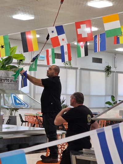 In a brightly lit indoor space, possibly a dining hall or community center in Be'er Sheva, Israel, two men are present. The man in the foreground, wearing a black polo shirt and dark pants, is actively engaged in hanging a string of small national flags from a wire suspended across the room. He holds a green flag and a blue and yellow flag in his hands as he reaches upwards. To his right, another man in a black t-shirt with a circular emblem on the back is seated, looking towards the left of the frame. The flags suggest a multinational or cultural event. Plants are visible in grey planters, and a row of orange chairs and tables are in the background, indicating a space designed for people to gather. The walls are white and feature blinds or screens.