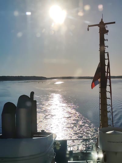 The image depicts a maritime scene, presumably near Edoma, Russia, viewed from a vessel at sea or on a large body of water during either sunrise or sunset. The sun, a brilliant white orb, dominates the upper left quadrant of the clear, light blue sky, creating significant lens flare and bokeh effects. Its intense light reflects across the water surface, forming a shimmering, sparkling path that extends towards the distant horizon.

In the foreground on the left, a portion of a white, enclosed lifeboat or emergency capsule is visible, with two dark, vertically oriented smokestacks or ventilation shafts positioned behind it. On the right side of the vessel, a tall, dark, utilitarian mast-like structure rises, equipped with several ladder-like rungs and various pieces of equipment at its summit, including a horizontal cross-arm. A tricolored flag, featuring red, blue, and another indistinct color, hangs partially unfurled from this mast. Below the mast on the right, part of the ship's railing is visible, and a white, cylindrical container or housing is present, bearing the Cyrillic inscription "АККУМУЛЯТОР" (AKKUMULYATOR), meaning "BATTERY" or "ACCUMULATOR."

The midground features a wide expanse of water with ripples and glinting sunlight. In the far distance, a dark, low-lying landmass, likely forested, stretches across the horizon, separating the water from the sky. Small, indistinct dark shapes are faintly visible on the water in the mid-distance, too vague to identify definitively as people or objects. No clearly discernible individuals are present in the scene. The overall environment suggests calm waters and clear weather conditions.
