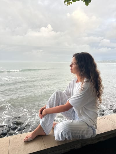 The image captures a woman in Pekutatan, Indonesia, kneeling on a tiled surface bordering a rocky shoreline where ocean waves break. She is dressed in a white t-shirt and light-striped trousers. Her long, curly brown hair cascades down her back as she leans forward, gazing downward with her hands clasped in front of her. The background features a grey, overcast sky meeting the horizon of a pale, rippling ocean. A small patch of green foliage is visible in the upper left corner.