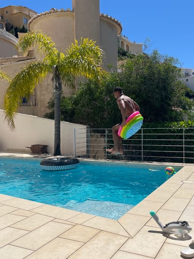 The image captures a man diving into a swimming pool in what appears to be a residential area of el Poble Nou de Benitatxell / Benitachell, Spain. The man, who is shirtless and wearing swim trunks, is in mid-air, with a colorful inflatable ring around his waist. He is positioned to enter the pool headfirst.
The pool is rectangular and filled with blue water. A black inflatable tire is floating in the water. A snorkel and diving mask are positioned on the deck of the pool. On the pool deck, the tiles are arranged in a herringbone pattern.
Behind the man, there is a white railing and dense green foliage. In the background, there are buildings, likely residential homes. The sky is clear and blue. A tall palm tree is visible to the left of the frame. The image appears to have been taken during daylight hours.