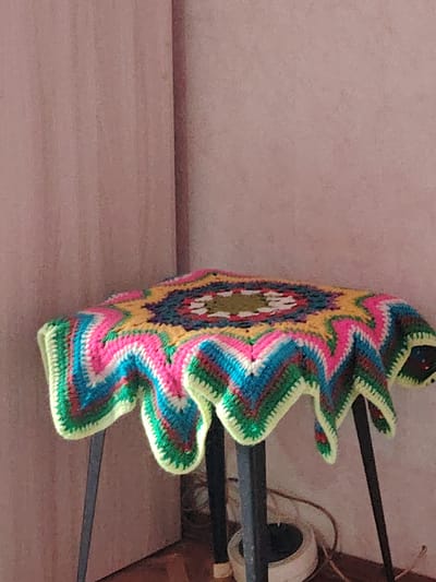 A vibrant, star-shaped crocheted blanket or mat is draped over a dark-legged stool or small table, positioned in the corner of a room in Yanaul, Russia. The crocheted item is a riot of color, featuring concentric patterns in bright pink, yellow, teal, blue, white, green, and a deeper shade like maroon or dark green. It has a distinctive zigzag or chevron design radiating outwards from the center, culminating in a wavy, scalloped edge bordered in bright lime green.
The stool or table supporting the blanket has slender, dark-colored legs, likely metal, with three visible in the frame. The background consists of two walls forming a corner. The wall on the left and center has a light pinkish-brown or mauve hue with a subtle vertical striation texture. The wall on the right appears to be a slightly different shade or texture of the same color, creating a distinct corner. The floor in the lower part of the image is a darker brown, possibly wood or carpet. On the floor near the stool's legs, a white circular object, which appears to be a base or electrical component, is visible, along with some thin cables or wires.
The scene is an indoor, still life arrangement, with no activity taking place. The lighting is ambient, without strong shadows to indicate a specific time of day. No people are present, and no text is visible in the image. The overall impression is one of handmade craft and a cozy indoor setting.