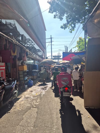 A narrow street scene in Parañaque, Philippines. The street is lined with buildings, some with corrugated metal roofs. Laundry hangs from the buildings on the left. There are people gathered under an awning further down the street, partially obscured by a red, white and blue flag. A motorcycle with a pink food delivery box is parked on the right side of the street. The motorcycle's license plate reads "727 XSO". Trash and debris are scattered along the street. Power lines crisscross overhead. Another motorcycle is partially visible in the foreground.