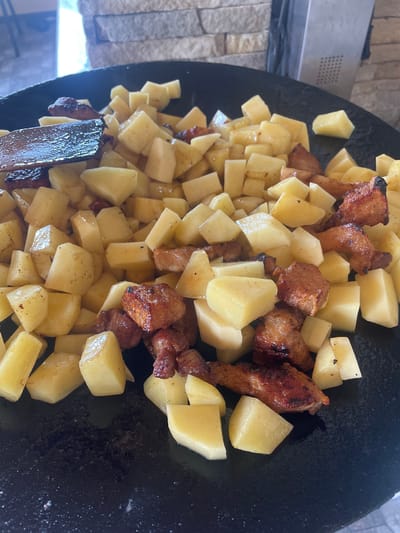 The image displays a close-up, overhead perspective of a large, dark, circular metal griddle or pan containing food actively cooking. Numerous irregularly diced potatoes, predominantly light yellow to off-white, are spread across the surface, mixed with browned pieces of meat, consistent with pork or chicken, exhibiting caramelization and dark seared edges. A dark wooden cooking utensil, possibly a spatula, is visible on the left side of the griddle, resting among the food. The background on the right features a dry-stacked stone wall constructed from rectangular, textured blocks in shades of grey and beige. Adjacent to this wall, a dark, vertical metallic element with a grid-like perforated pattern is partially visible. The upper-left background is out of focus, revealing ambient light and indistinct structural elements. The scene depicts the preparation of a meal in an outdoor or semi-outdoor environment located in Petrivka, Ukraine. No individuals are present in the frame.