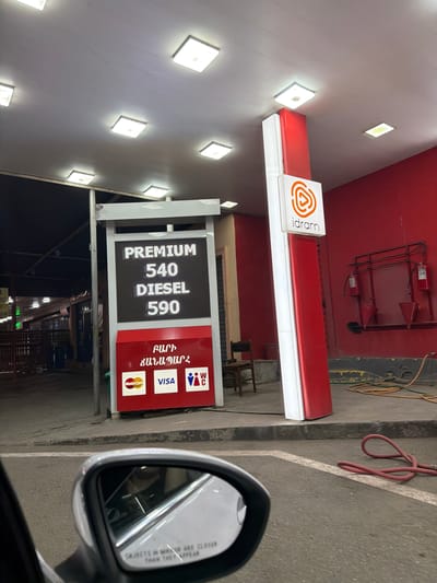 The image shows a gas station in Parakar, Armenia. The gas station has a red and white structure with an "idram" logo at the top. A digital display shows fuel prices, with "PREMIUM 540" and "DIESEL 590" visible. Below the display, there are logos for MasterCard, VISA, and a WC sign. A red hose is coiled on the ground. The scene appears to be captured at night, with overhead lighting illuminating the area. The perspective is from inside a vehicle, looking out through the side mirror, which reflects a portion of the interior and the road. There are no people visible in the main frame of the image.