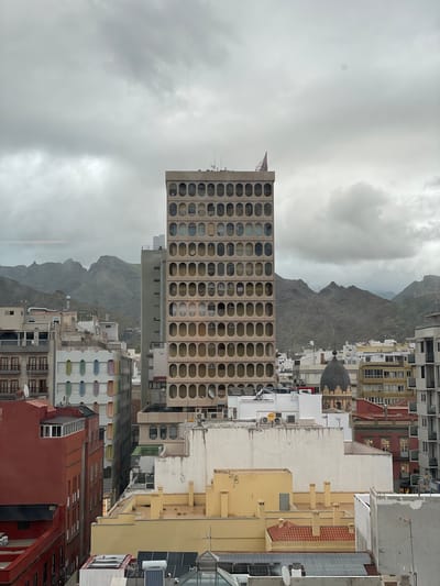 The image presents a dense urban cityscape against a backdrop of mountains and an overcast sky. A prominent, multi-story building, likely a hotel or large residential block, dominates the mid-ground on the left, characterized by a striking, repetitive facade of oval-shaped windows or openings. The building is light-colored, possibly concrete or a similar material.
Beyond this building, and stretching across the background, are rugged, rocky mountains, their peaks partially obscured by the heavy, grey clouds, indicating an overcast day.
In the foreground and to the right, a close-packed arrangement of various buildings fills the frame. These buildings feature diverse architectural styles and colors, including yellow, white, red, and beige structures. Many rooftops are visible, some equipped with solar panels, antennas, satellite dishes, and water tanks. One rooftop clearly shows the brand name "JUNKERS" on a white cylindrical tank, likely a water heater. The overall impression is one of a busy, established urban environment. There is no visible human activity, giving the scene a quiet, almost still quality despite its density.