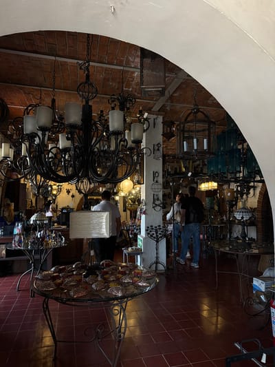 The image displays the interior of a retail establishment specializing in lighting fixtures and home decor, located in Tlaquepaque, Mexico. The high ceiling features exposed red brick barrel vaulting, partially framed by white painted architectural arches. The floor is uniformly covered with dark red square ceramic tiles.
Numerous chandeliers, lanterns, and lamps of various designs are suspended from the ceiling and displayed on tables. A large black wrought-iron chandelier with multiple arms and frosted glass shades dominates the left-center foreground. Other fixtures visible include cage-style lanterns and chandeliers featuring green-tinted glass elements on the right side.
Two circular glass-top tables with wrought-iron bases are prominent in the foreground. The table on the left displays multiple small, colorful, possibly stained-glass butterfly-shaped items. The central table holds an arrangement of numerous small, metallic, textured, domed decorative objects, interspersed with smaller yellow items.
Four individuals are present. In the center-right, an adult male wearing a light-colored polo shirt and dark pants is carrying a large, rectangular lampshade wrapped in clear plastic. To his right, an adult male with a dark top and blue jeans stands alongside an adult female in a light top and blue jeans; both are observing merchandise. In the mid-left background, an adult female with dark hair is partially visible behind a counter. Further to the left, a person with reddish-blonde hair and glasses is visible behind a service counter.
A red fire extinguisher is situated on the floor near the right edge of the frame, next to a white box labeled "TOALLA INSTITUCIONAL." The scene is lit by the numerous display fixtures, casting reflections across the tiled floor.