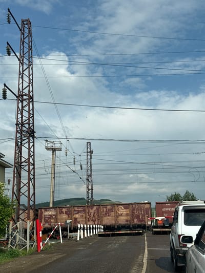 A railway crossing is depicted in Zykova, Russia, with multiple train cars stopped on the tracks. Two large, rust-colored metal towers supporting overhead electrical lines are prominent in the foreground and midground. The sky is a mix of blue with scattered white clouds. A red and white striped barrier gate is lowered across the road, indicating the train's passage. A white SUV is stopped at the crossing. The road appears to be paved with asphalt. The surrounding environment shows greenery and distant hills.