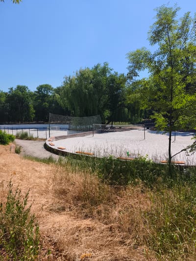 The image depicts a bright, sunny day with a clear blue sky, overlooking a sandy sports court which appears to be a volleyball or beach tennis court. The court is surrounded by a low, curved wall made of wood or composite material, with several orange benches visible along its edge. A white net is stretched across the court, supported by poles. Beyond the court, a perimeter fence encloses the area, and further back, more trees and greenery are visible, suggesting a park or recreational area. In the foreground, dry, yellowed grass and some green foliage create a natural border. The location is identified as Sokolivske, Ukraine. There are no people clearly visible in the immediate foreground or on the court itself.