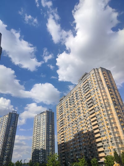 The image shows a view of modern apartment buildings against a blue sky with white clouds. Three tall, multi-story buildings are visible. The building on the far right is the largest and appears to be made of brick and has balconies with yellow accents. To its left are two slightly shorter buildings, one with a darker facade. The buildings are residential, as indicated by the numerous windows and balconies. Trees are visible at the bottom of the image, suggesting a green space or park area. The scene depicts an urban environment, likely a residential district. The location in Labinsk, Russia is not indicated by any visual cues in the image itself.