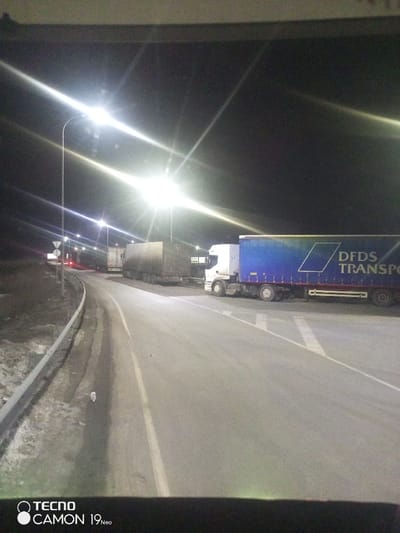 A nocturnal scene depicts a roadway in Pridorozhnyi, Russia, illuminated by multiple bright streetlights on tall poles, generating significant lens flare. The image is captured from a vehicle's interior, showing a dark dashboard at the bottom. Several semi-trailer trucks are visible on or adjacent to the asphalt road. In the foreground, on the right, a white-cabbed truck with a blue trailer bearing the white "DFDS TRANSPO" (likely "DFDS TRANSPORT") branding and a geometric logo is prominently positioned, seemingly stationary or turning. Behind it and further left, other trucks, mostly dark or covered in dirt, are observed. A guardrail runs along the left edge of the road, paralleling a barren, dry embankment. Multiple indistinct road signs on poles are visible near the guardrail. The road surface appears dry, and the sky is dark. No individuals are discernible.