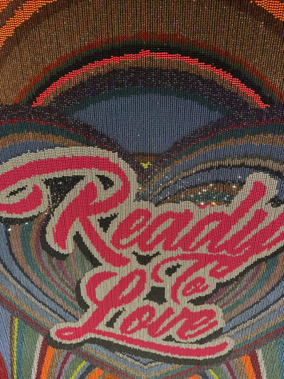 The image is a close-up, overhead shot of a colorful, beaded tapestry or artwork featuring the words "Ready for Love" in large, stylized pink lettering with white and black outlines. The lettering is centered within concentric circles of various colors, including blue, gray, green, brown, and orange, all formed by small beads. The beads themselves appear to have a slight shimmer or sparkle. This artwork is part of the Swarovksi Kristallwelten (Crystal Worlds) in Wattens, Austria, specifically located in the "Great Hall" where it is a permanent installation. There are no people or actions depicted in the image.