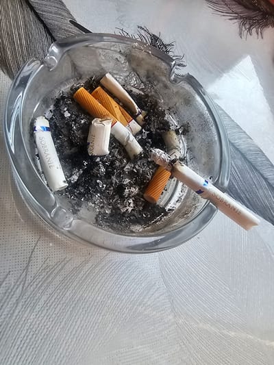 An ashtray filled with cigarette butts and ash sits on a textured white surface. The ashtray is made of clear glass and contains several Sobranie cigarette butts, some with their original orange filter tips visible. One cigarette butt is lit and rests across the edge of the ashtray, with ash falling onto the other contents. The background features a pattern resembling feathers. The scene is set indoors, likely in a residential area of Таёжное, Russia. There are no people visible in the image, and no actions are being directly observed other than the presence of the extinguished and one partially burnt cigarette.