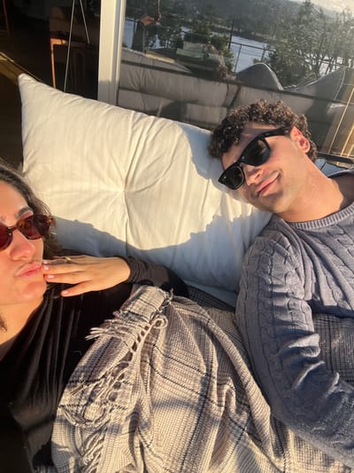 Two people are lying side-by-side on a white cushion. One person, on the left, is wearing a grey cable knit sweater and black sunglasses. The other person, on the right, is wearing a black shirt and brown sunglasses, with a beige and black plaid shawl partially covering them. The scene appears to be outdoors, possibly on a deck or patio, as suggested by a nearby window reflecting foliage. The location is Gondarém, Portugal.
