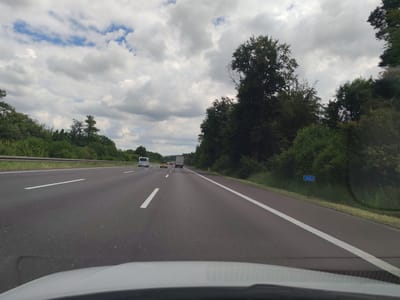 The image captures a daytime scene from inside a vehicle driving on a multi-lane highway, likely an autobahn in Germany. The sky overhead is mostly covered with large, fluffy white and grey clouds, with glimpses of blue sky visible between them.

The asphalt road dominates the lower and middle portions of the frame, showing several lanes marked by white dashed lines and a solid white line on the right shoulder. The vehicle from which the photo is taken has its dashboard or hood visible at the bottom of the image.

Ahead, several vehicles are seen in different lanes: a white cargo van in the far left lane, a bright yellow car in the middle lane, and a large truck further in the distance. Lush green trees and dense foliage line both sides of the highway, extending into the background. A metal guard rail runs along the left side of the road.

On the right side, a blue rectangular sign is visible on the embankment, displaying the white text "143.5". The weather appears overcast but bright, characteristic of a cloudy day. The setting is rural, surrounded by natural vegetation.