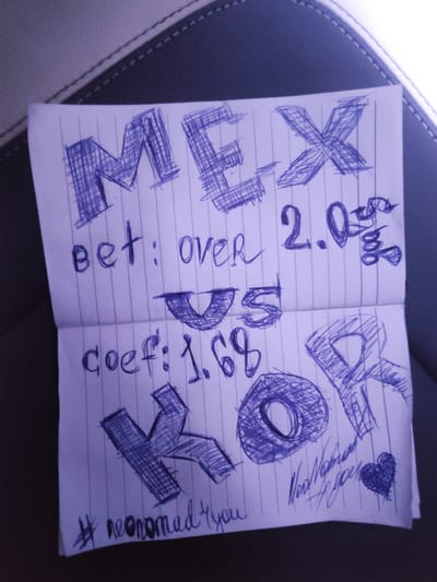 The image shows a close-up view of an open notebook with a handwritten message and drawings. The notebook is placed on a dark, textured surface, possibly the interior of a vehicle. The notebook has lined paper and is written on with a blue pen.
The top of the notebook displays the word "MEX" in large, stylized letters. Below this, the text reads "bet: over 2.05". There are also drawings of what appear to be stylized letters "VS" and "KOR". Further down, the text "coef: 1.68" is visible, followed by more stylized letters, with "K" and "O" being most prominent. At the bottom of the notebook, there is a hashtag "#neonamadryou" and a handwritten signature that reads "Neo Nomad you", accompanied by a small drawing of a heart.
The location in the city of Xuân Long, Vietnam, is not discernible from the provided image.