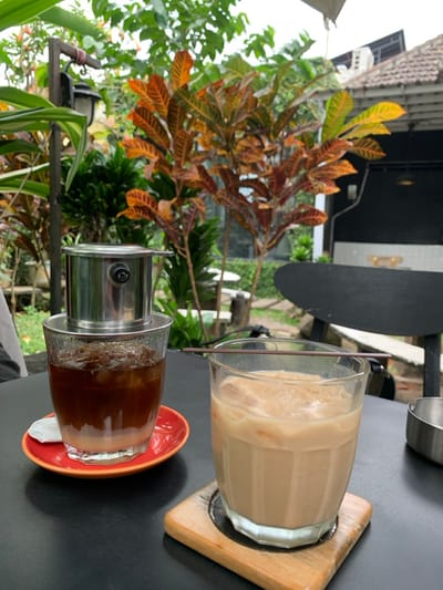 The scene depicts an outdoor cafe setting in Kota Batu, Indonesia. In the immediate foreground, two iced beverages are positioned on a dark, flat table surface. The left glass contains a layered drink with a dark liquid over a lighter opaque liquid, ice cubes, and is topped with a stainless steel Vietnamese phin coffee filter. This glass rests on a red ceramic saucer, with a small white paper napkin visible beneath it. To its right, a second clear glass holds a light brown, milky iced beverage with ice cubes, and a brown plastic straw is laid horizontally across its rim. This glass is situated on a square wooden coaster with a circular metal insert. A partially visible small silver metal container is present on the far right of the table. The background features dense, varied vegetation, including plants with prominent orange and red patterned leaves, suggesting a lush garden environment. A dark wooden post with a black lantern is visible on the left side amidst the greenery. On the right, a dark building structure with a terracotta tile roof is present. Several white outdoor tables or stools are discernible in the mid-ground among the foliage. The sky above is bright, indicating daytime conditions. No individuals are present in the frame.