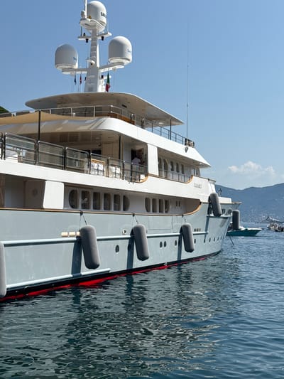 The image shows a large, grey luxury yacht docked in a body of water, likely a harbor, with a mountainous coastline in the background under a clear blue sky. The yacht has multiple decks, white railings, and large, rounded windows. Several grey, cylindrical fenders are attached to its side, suggesting it's docked or moored. The name "ELISABET" is visible in gold lettering on the side of the yacht.
In the foreground, several white satellite domes are mounted on a mast structure. The water is a deep blue with ripples reflecting the sunlight and the yacht's hull. The scene appears to be set during the daytime, with bright sunlight casting shadows. The overall impression is one of opulence and leisure, typical of a high-end yacht in a scenic location. The presence of mountains and clear skies hints at a pleasant, possibly warm weather day.