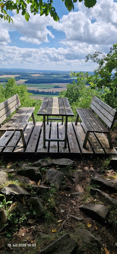 This image shows a wooden picnic table and benches with a scenic view of rolling hills and fields under a partly cloudy sky. The foreground is a rocky, wooded area. The date and time stamp "19. Juli 2026 14:33" is visible in the bottom left corner. The location is identified as Neustadt am Kulm, Germany, suggesting this is a viewpoint or rest area in a natural setting. There are no people present in the image.