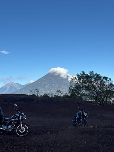 The scene depicts an outdoor volcanic landscape in San Vicente Pacaya, Guatemala, under a clear blue sky with minimal cloud cover. The foreground consists of dark, uneven, granular earth. Two motorcycles are stationary on this terrain. The motorcycle on the left is a dark red/maroon cruiser-style model with visible chrome elements, including the front wheel and headlight. A section of a person's lower left leg, covered by dark trousers, is visible at the extreme bottom-left, adjacent to the left motorcycle. The second motorcycle, on the right, is a darker, possibly black, sportier-style model, angled slightly towards the viewer. In the midground and background, multiple volcanoes are visible. The largest, centrally located, has a dense white cloud formation obscuring its summit and upper slopes, with greener vegetation discernible lower down. To the left of this primary volcano, two additional, more distant mountains or volcanoes are present, appearing lighter in tone due to atmospheric perspective. Sparse trees and shrubs are scattered across the landscape, particularly on the right and midground slopes.