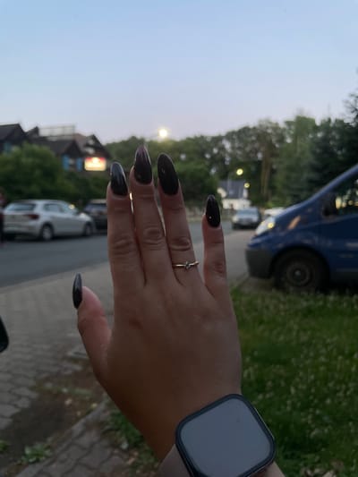 A person's hand with dark, elongated nails is positioned in the foreground of the image. The hand is adorned with a delicate silver ring on the ring finger. A smartwatch with a dark band and a square, dark screen is visible on the wrist. The background suggests an outdoor setting with a road, grass, and trees. The sky is a pale blue, indicating it is either early morning or late evening. In the distance, there are buildings and vehicles, including a blue van. The overall mood appears casual and relaxed. The image was taken in Bolesławiec, Poland.